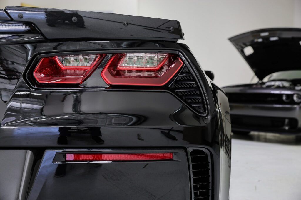 Black sports car's rear, red taillights, in garage with another car's open hood.