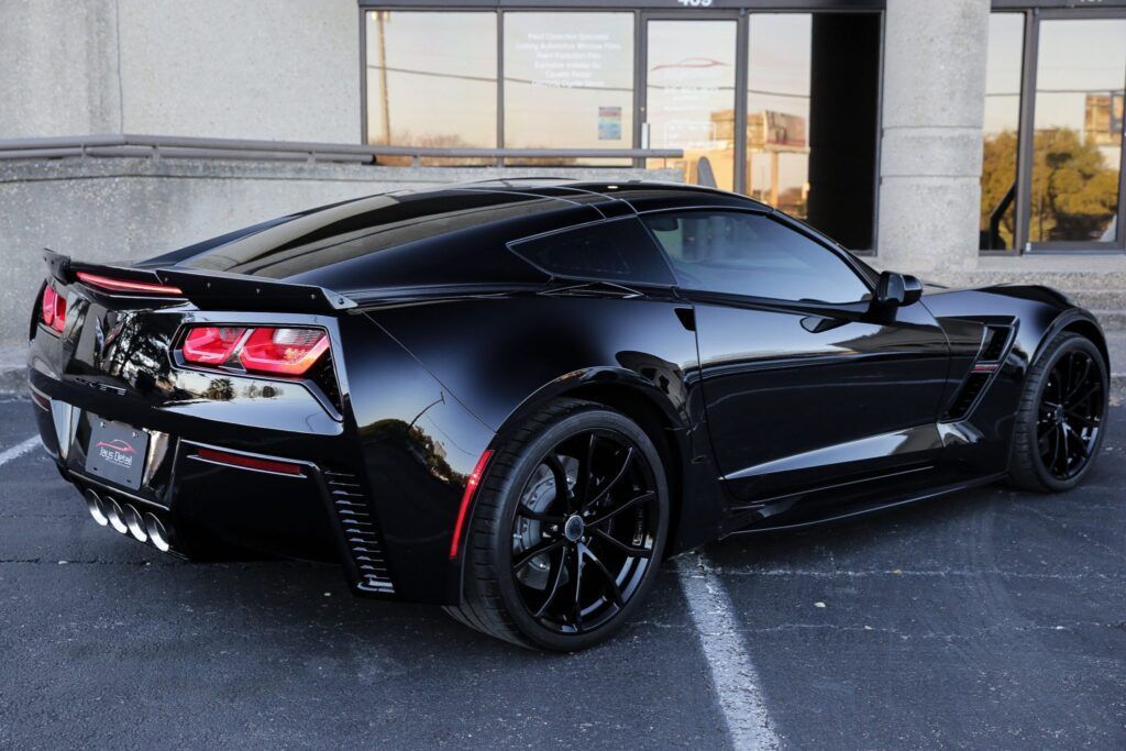 Black Corvette sports car parked in front of a building with glass doors, black wheels, and a rear spoiler.