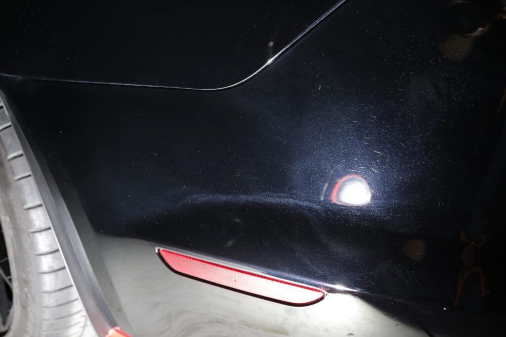 Black car bumper with swirl marks, a red reflector, and a tire visible.