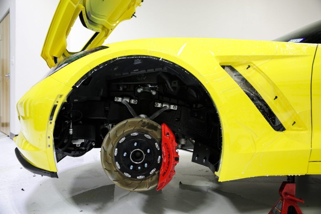 Yellow Corvette, front wheel removed, suspended on a jack, showing brake system. Hood open.