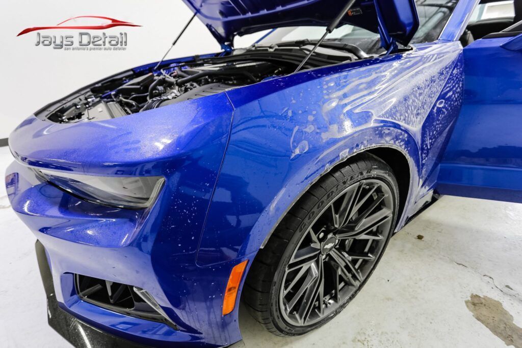 Blue Chevrolet Camaro being washed, hood and door open, spraying water on the front.