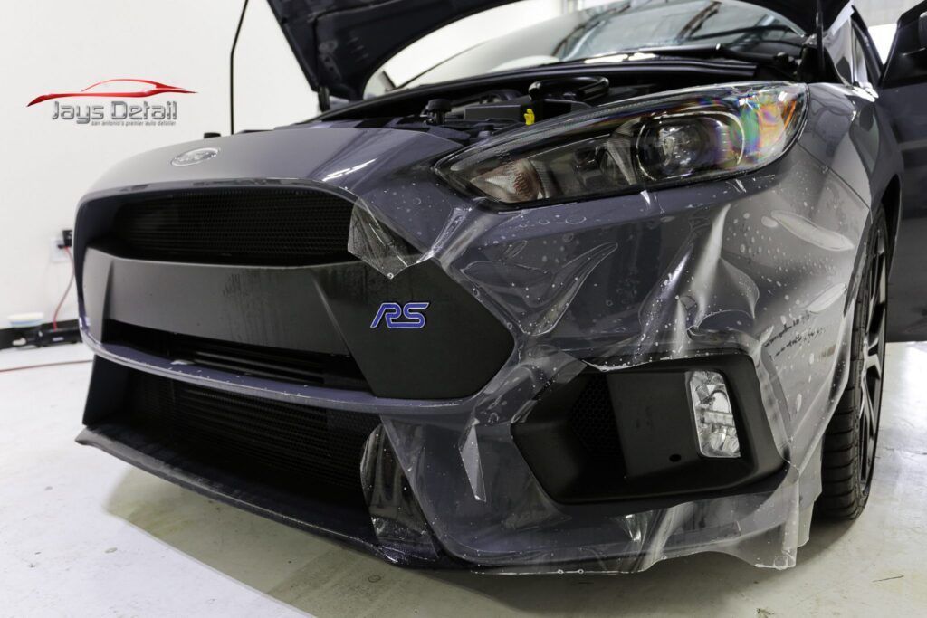 Gray Ford Focus with protective film on the front bumper, hood open, detailing shop setting.
