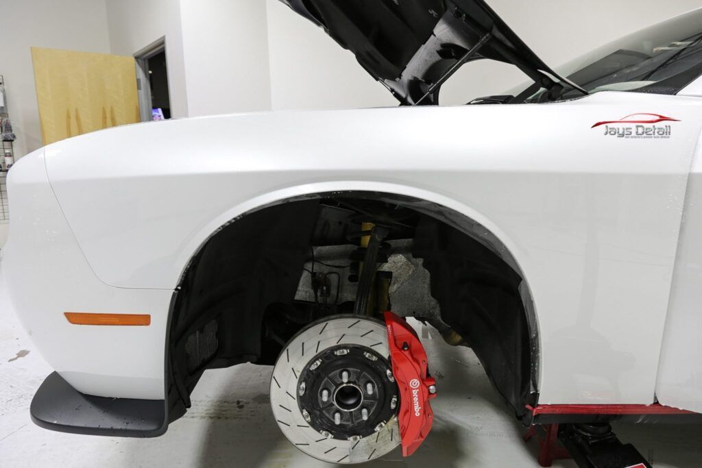 White car's front wheel, red brake caliper visible. Hood open, indoors.