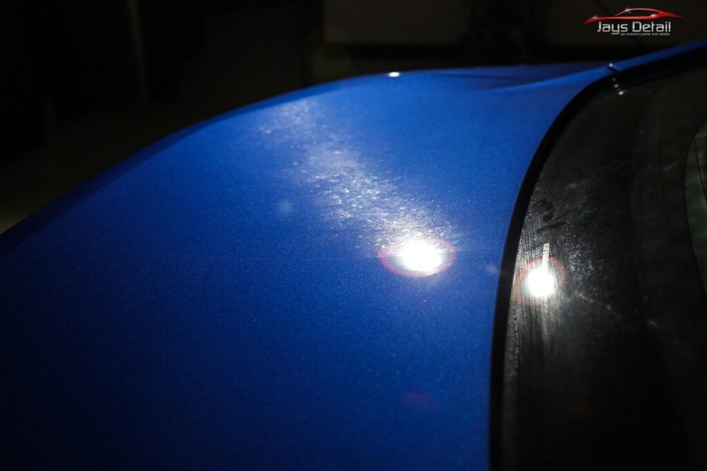 Blue car hood with faded, speckled paint. Reflection of a light source in the windshield.