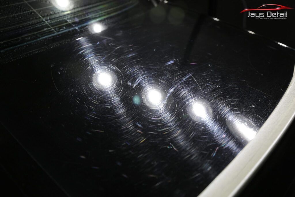 Black car hood surface with swirl marks reflecting bright lights.