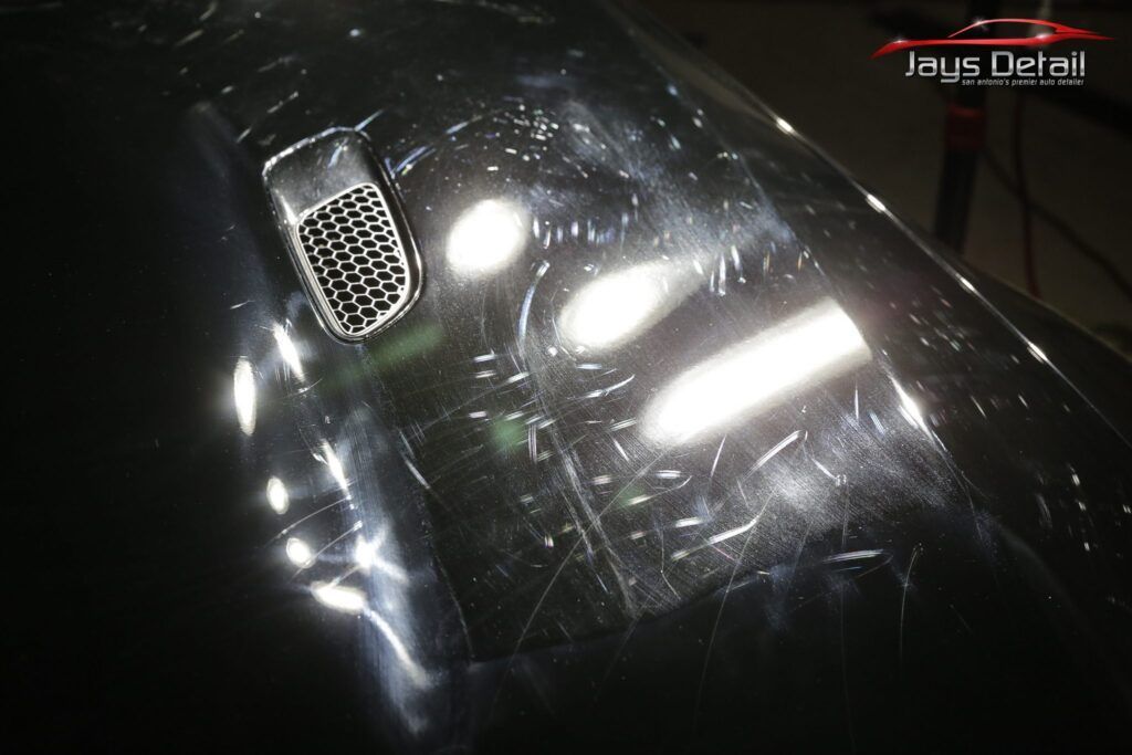 Close-up of a scratched black car hood with a vent visible; multiple light reflections.