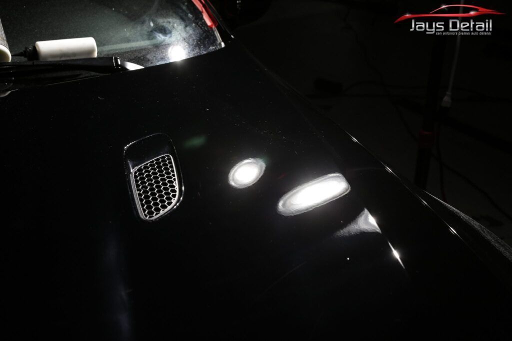 Black car hood reflecting light. Small vent visible. Detailing work in progress.