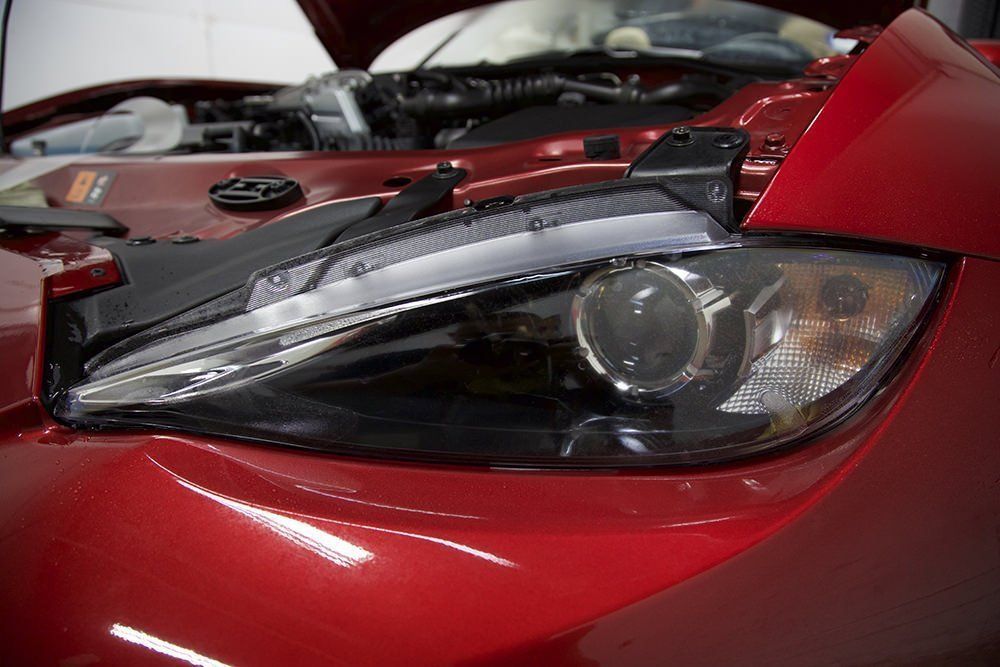 Red car with headlight removed, hood open in a garage.