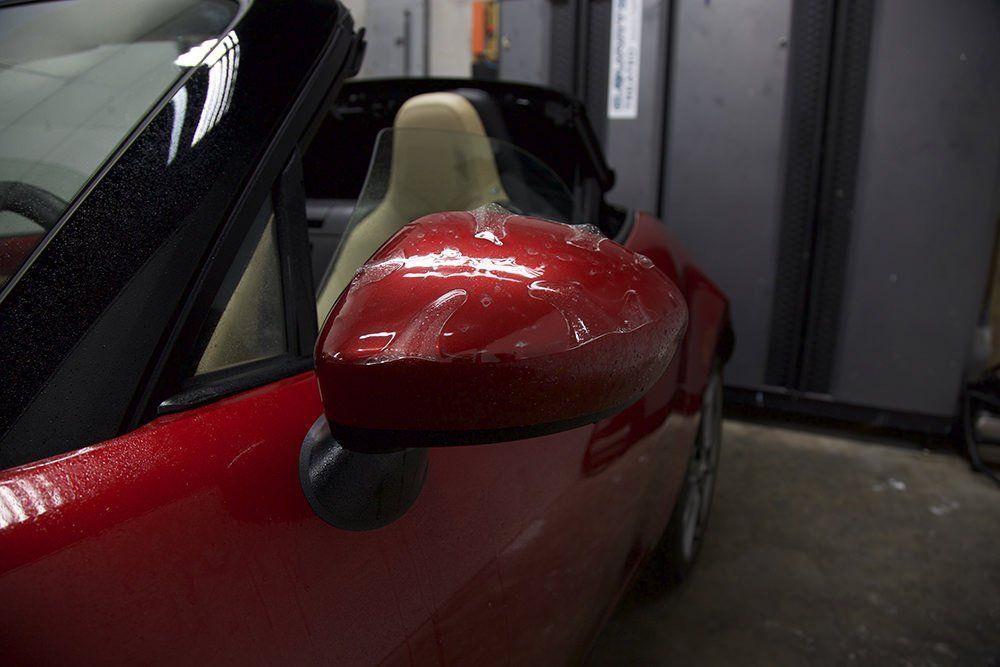 Red car's side mirror with a clear protective film, inside a garage.