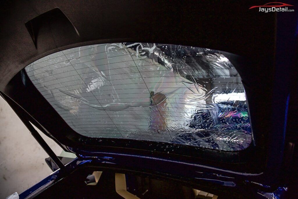 Tint being applied to a car's rear window; clear film and water visible. Inside car with open trunk.