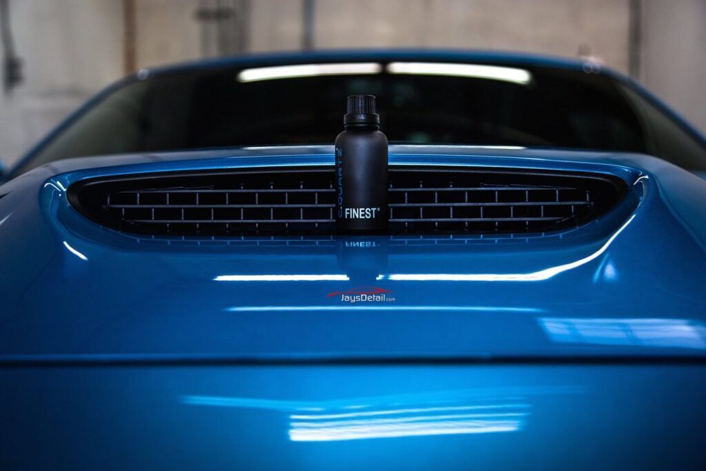Black spray bottle on a blue car hood with a vent, showing glossy paint and a garage setting.