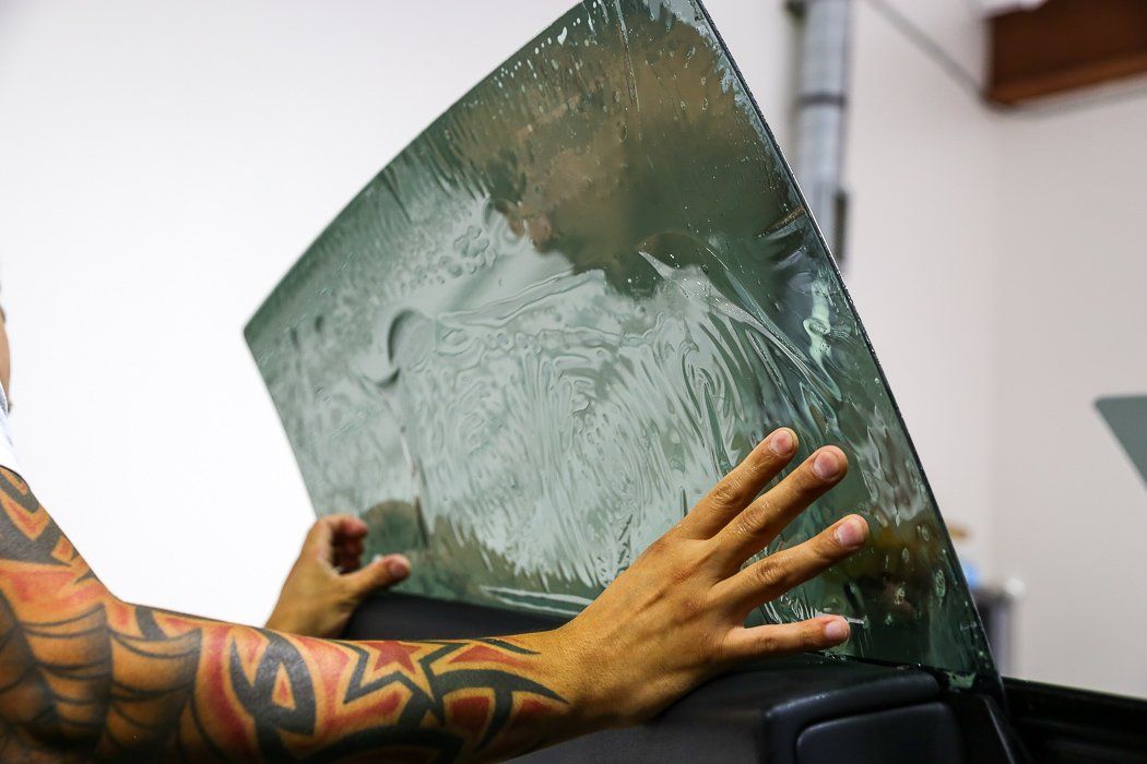 Person with tattoos applying window tint to a car window in a workshop.