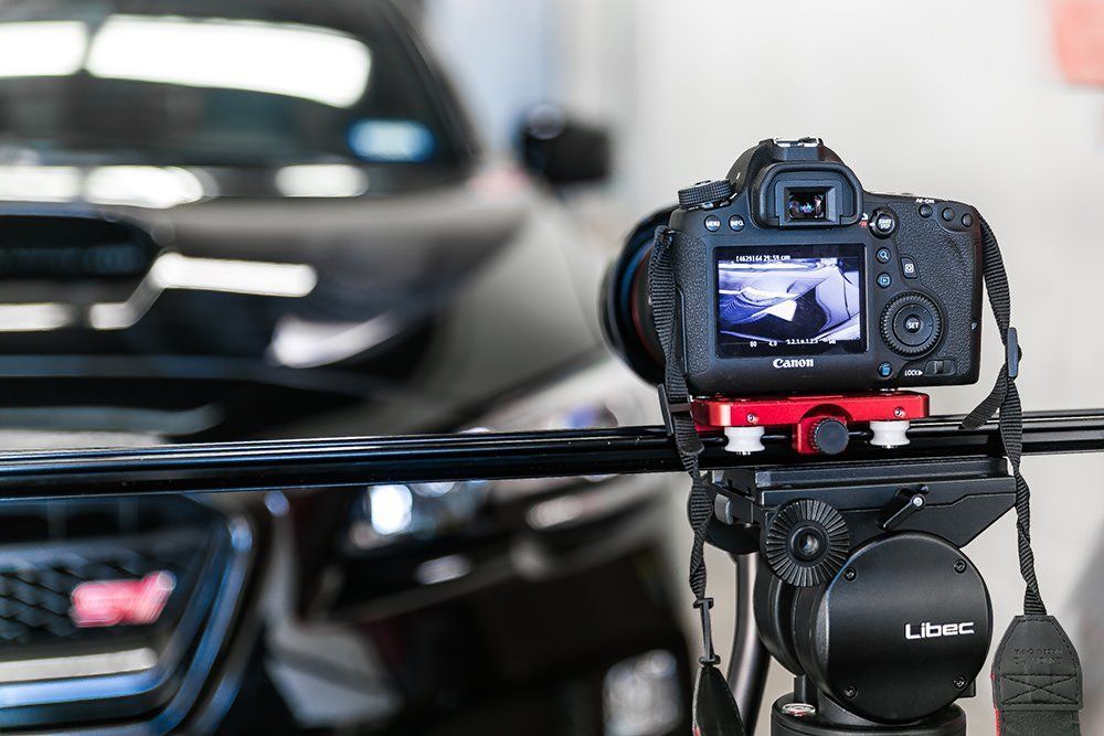 Camera on tripod filming a black car; close-up shot, red accents, automotive setting.