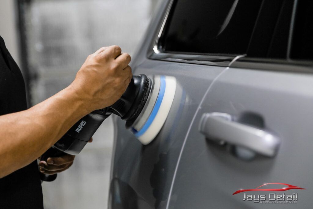 Person polishing a gray car door with a power buffer in a garage.