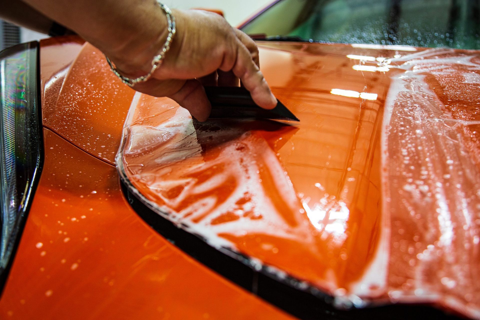 Hand using a squeegee to apply protective film to a bright orange car hood.