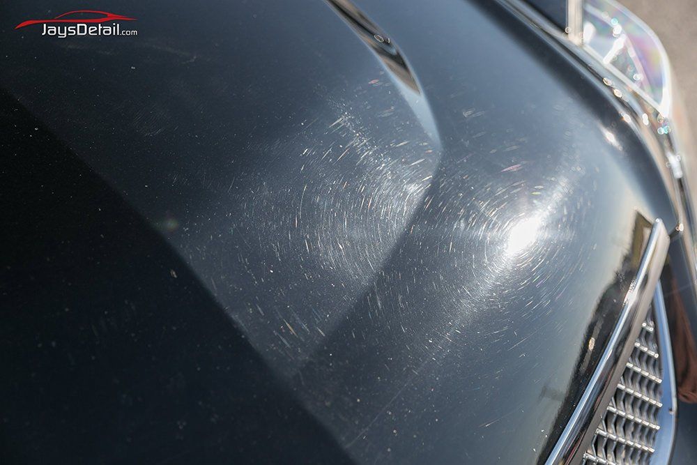 Close-up of a black car hood with numerous visible scratches reflecting sunlight.