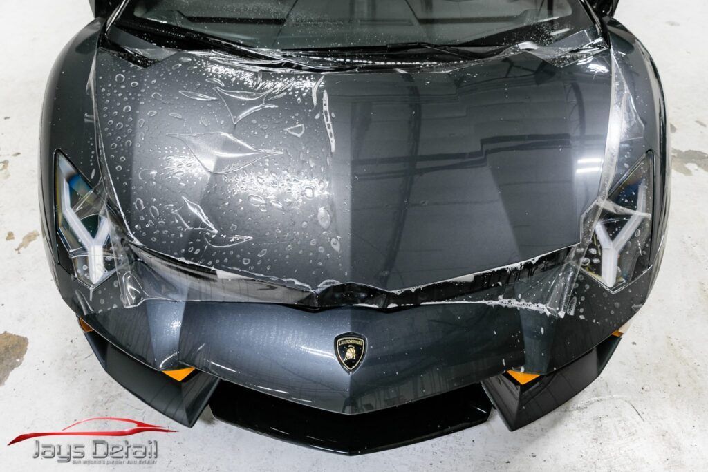 Lamborghini hood, partially covered in clear protective film being peeled off. Gray car, white floor, Jay's Detail logo.