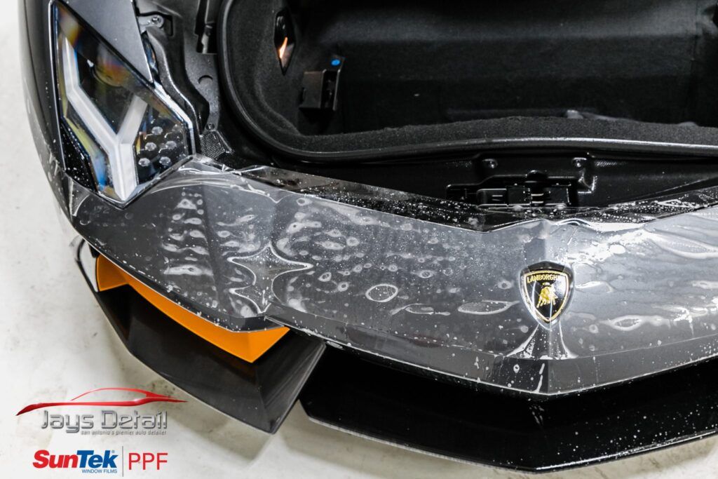 A black Lamborghini's front end with PPF installation, showing bubbles, a logo, and orange accents.