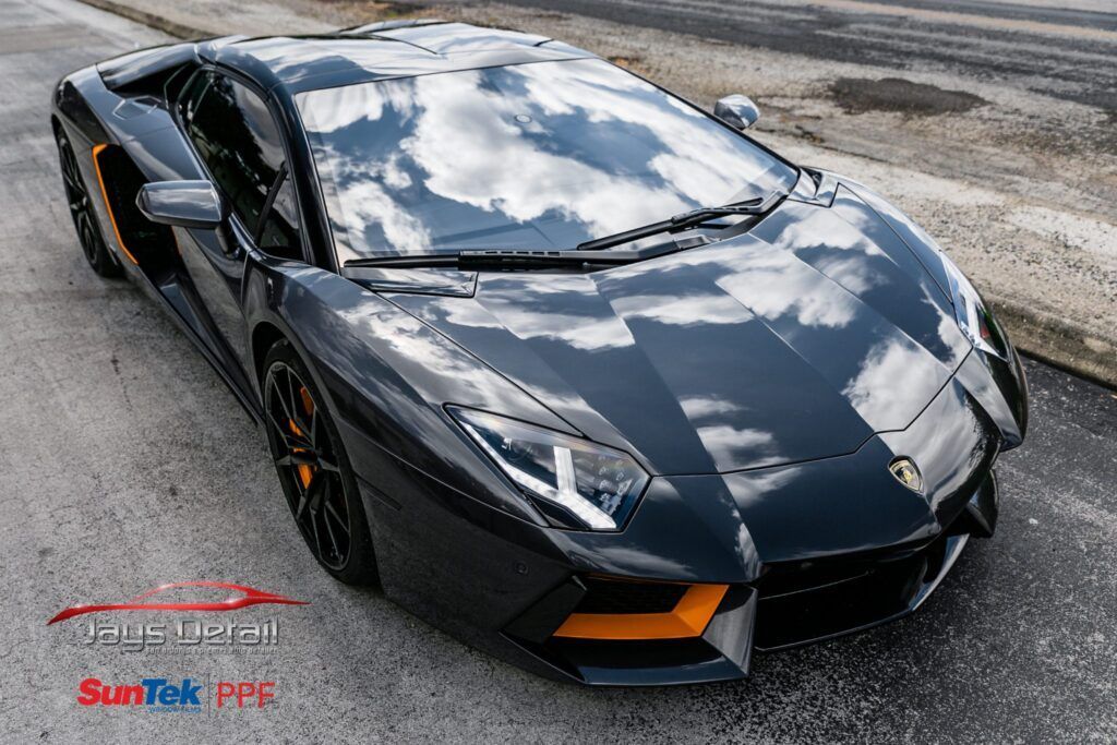 Black Lamborghini Aventador with orange accents parked on asphalt, reflecting the sky.