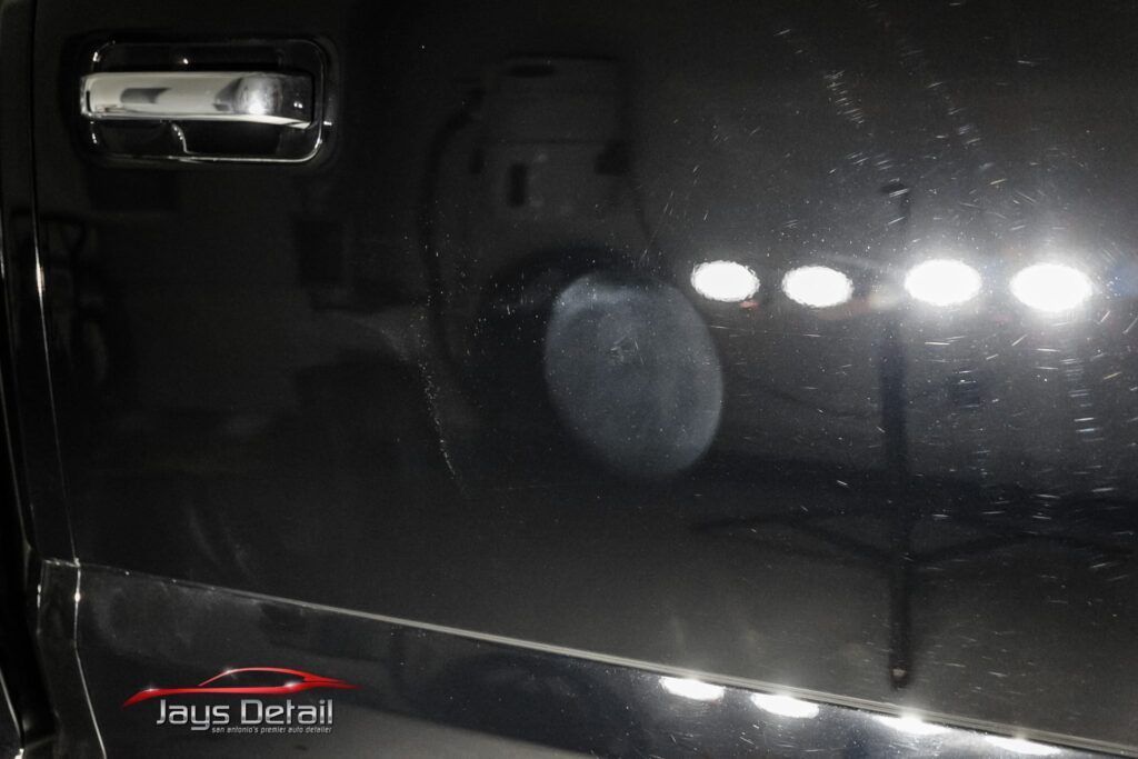 Black car door with swirl marks reflecting bright lights from detailing.