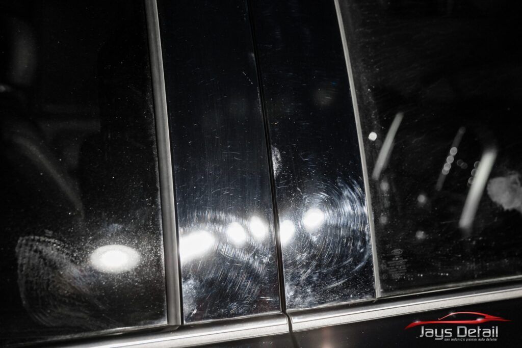 Scratched black car door frame reflecting light, showing swirl marks, inside a shop.