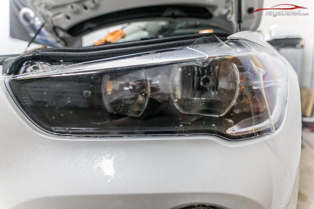 White car's headlight close-up; open hood; film applied; indoor setting.