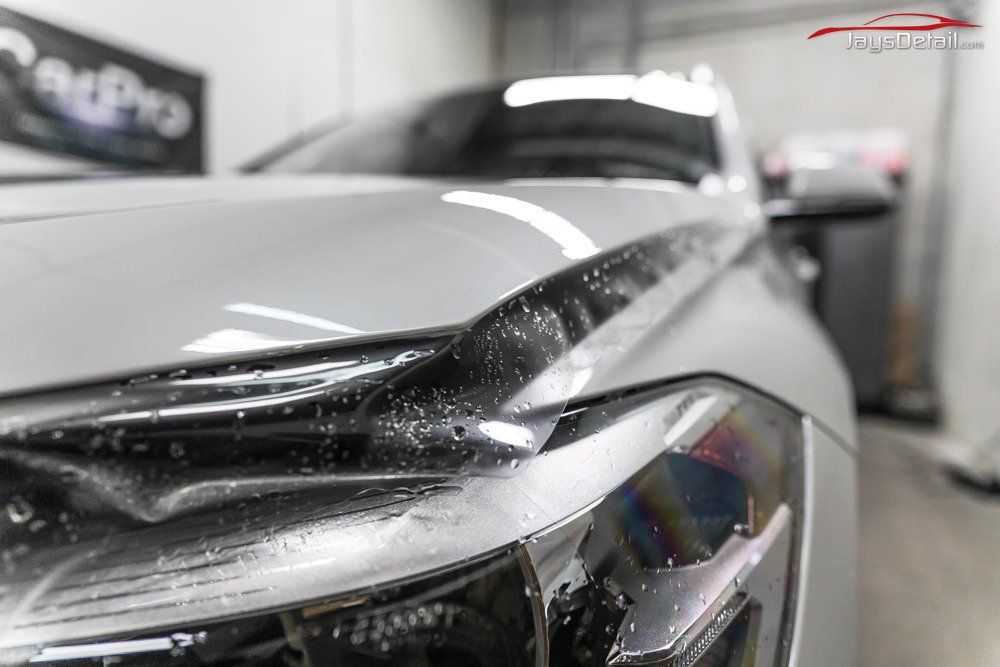 Close-up of a white car's headlight and hood with protective film applied. Inside a garage.