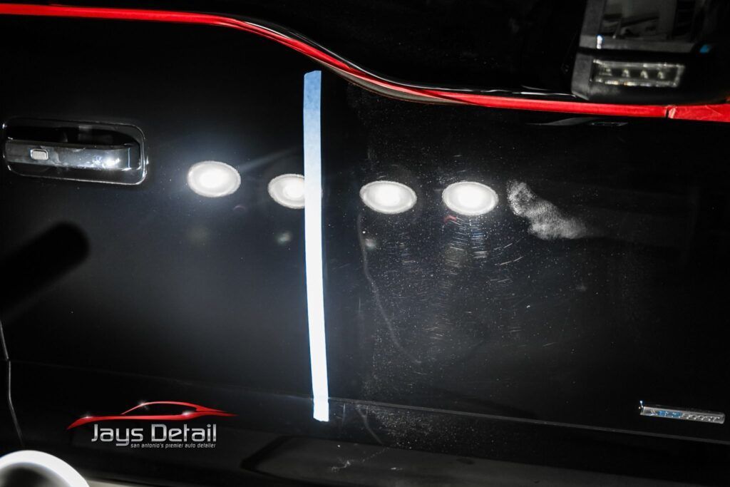 Black car door, one side polished, other side scratched, with tape dividing sections under spotlights.