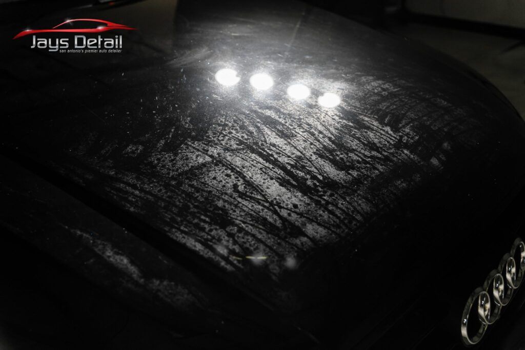 Black car hood with severe swirl marks, illuminated by overhead lights. Audi emblem visible.