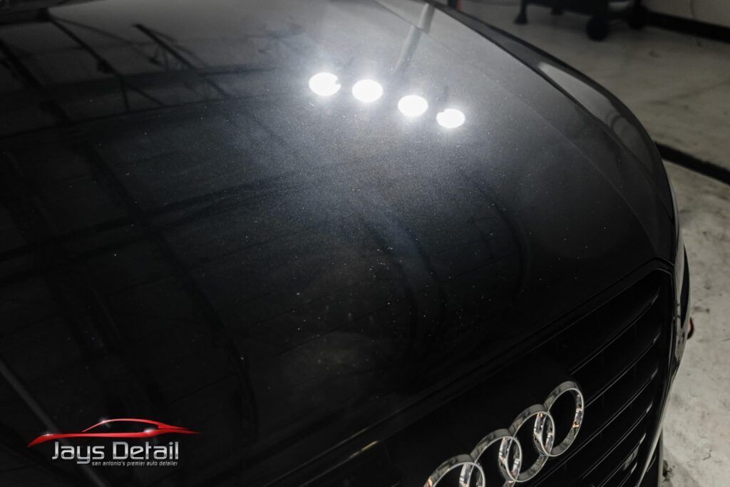 Black car hood with swirl marks under bright lights, showing an Audi emblem.