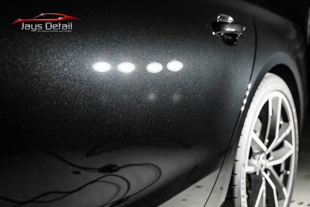 Black car door with light reflections, showing detailing work, and a wheel.