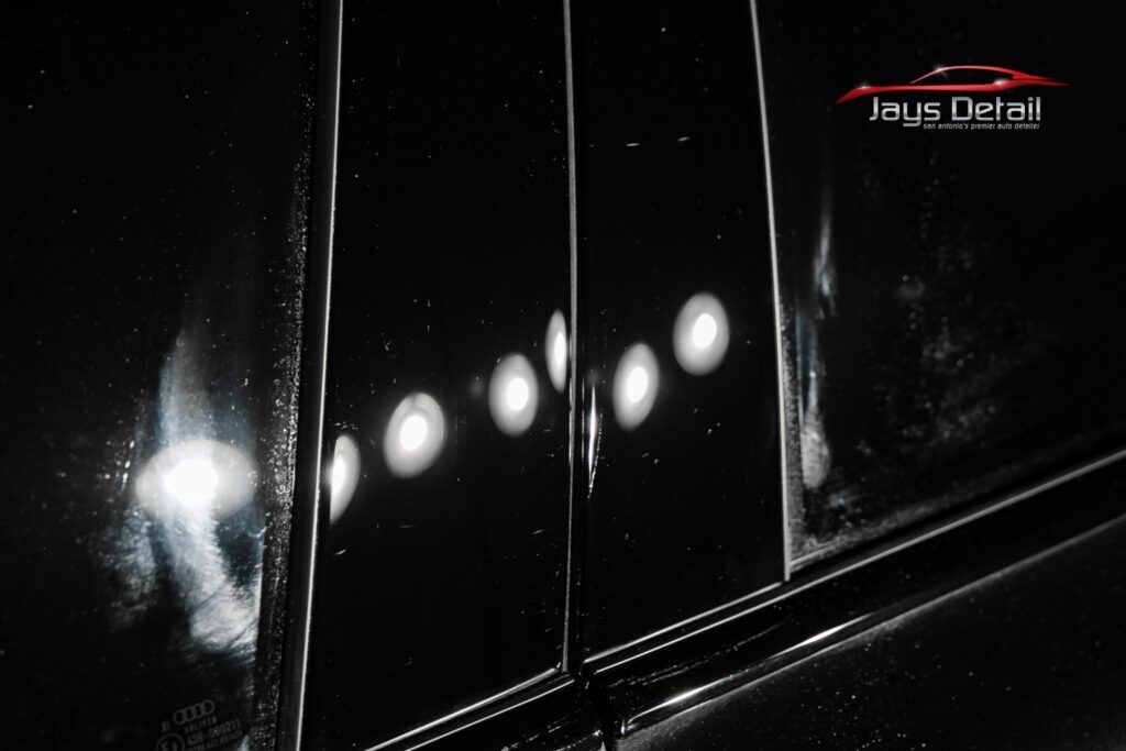 Black car door reflecting bright lights, showing scratches and swirls.