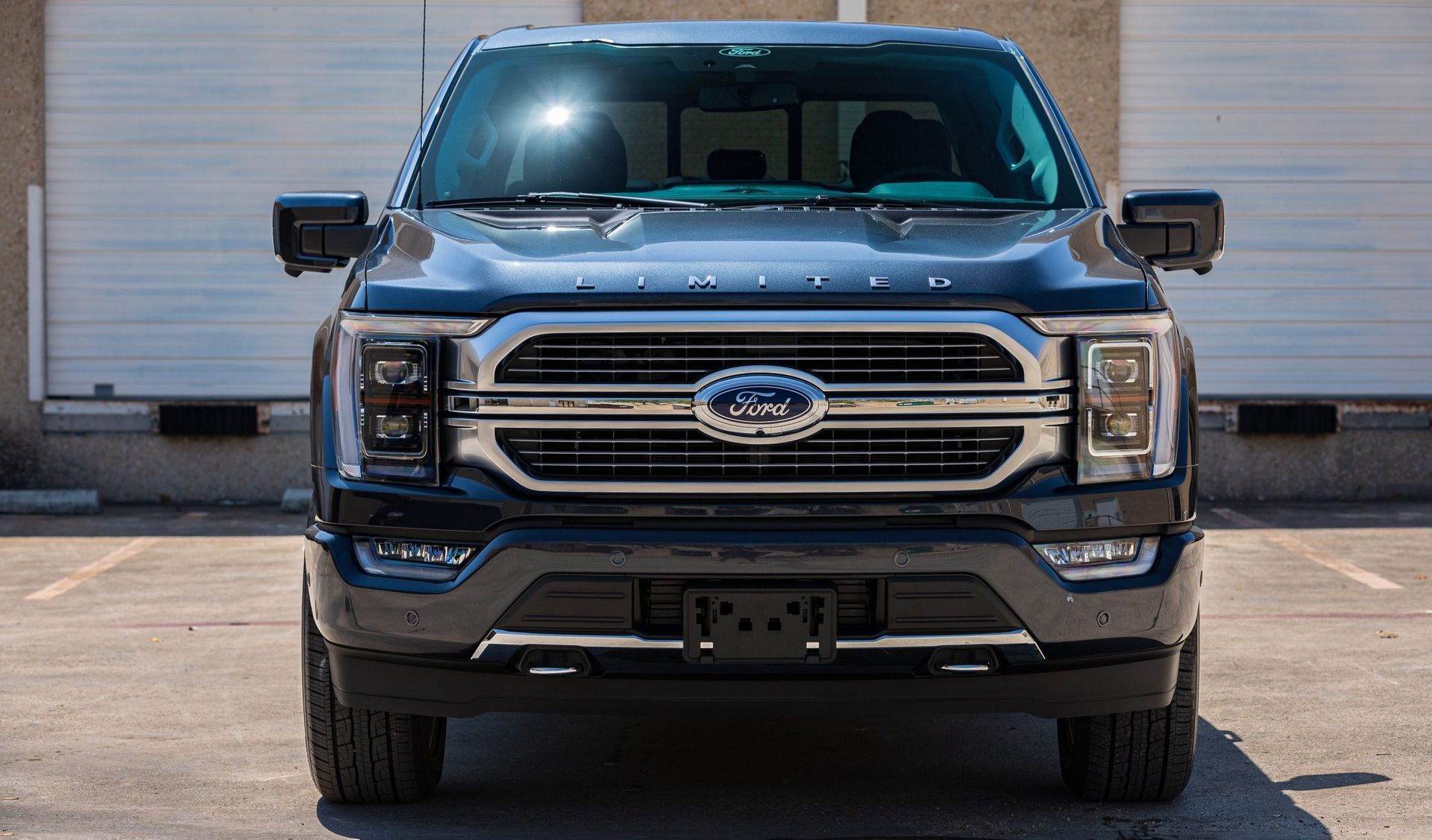 Ford F-150 truck, dark blue, front view. Parked in front of a tan building. Shiny chrome grille, sunny sky reflection.
