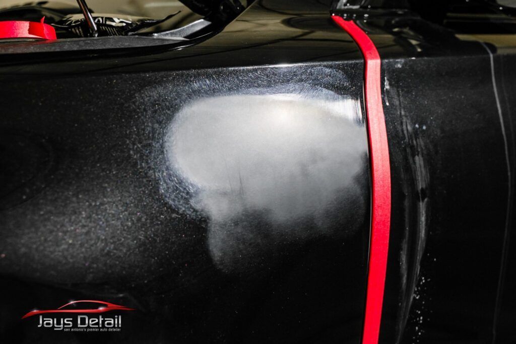 Black car panel with primer spot; red tape; detailer's work in progress.
