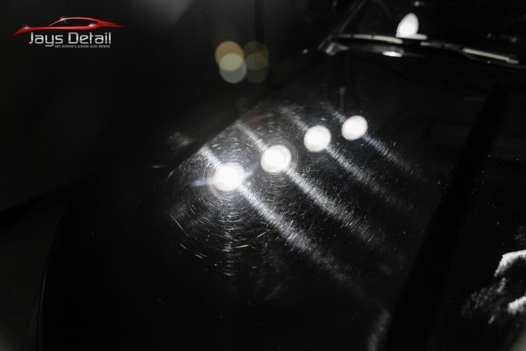 Black car hood with scratches, lit by overhead lights, reflecting streaks.