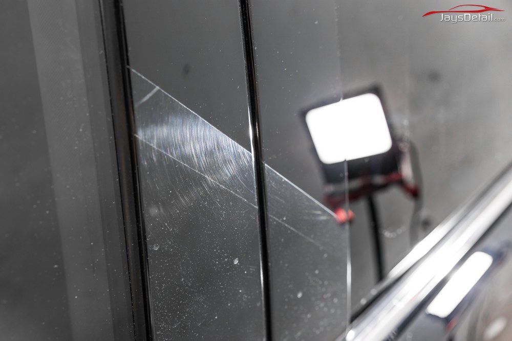 Scratched black car door panel, showing damage from abrasive contact, with reflection of light.