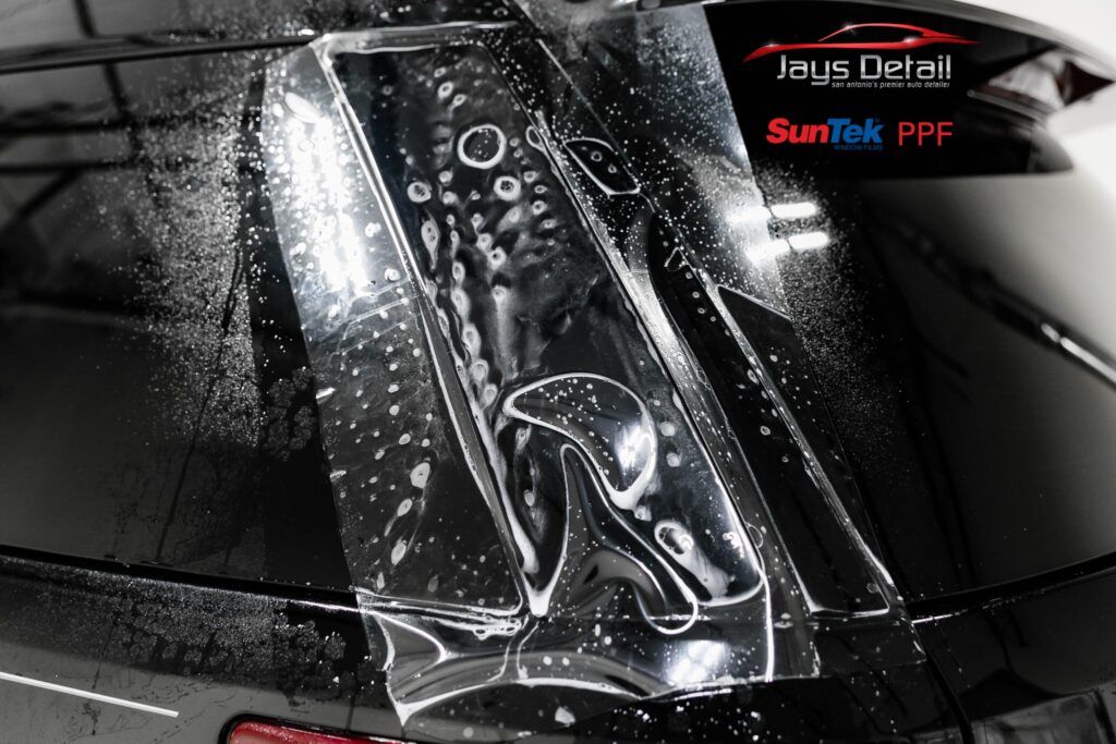 Clear protective film being applied to a black car's rear window, covered in soapy water.