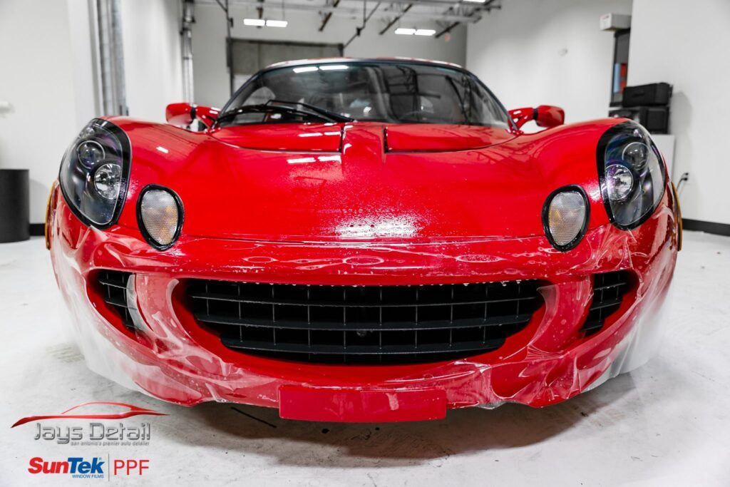 Red Lotus sports car covered in plastic wrap, likely for paint protection. Indoor setting.