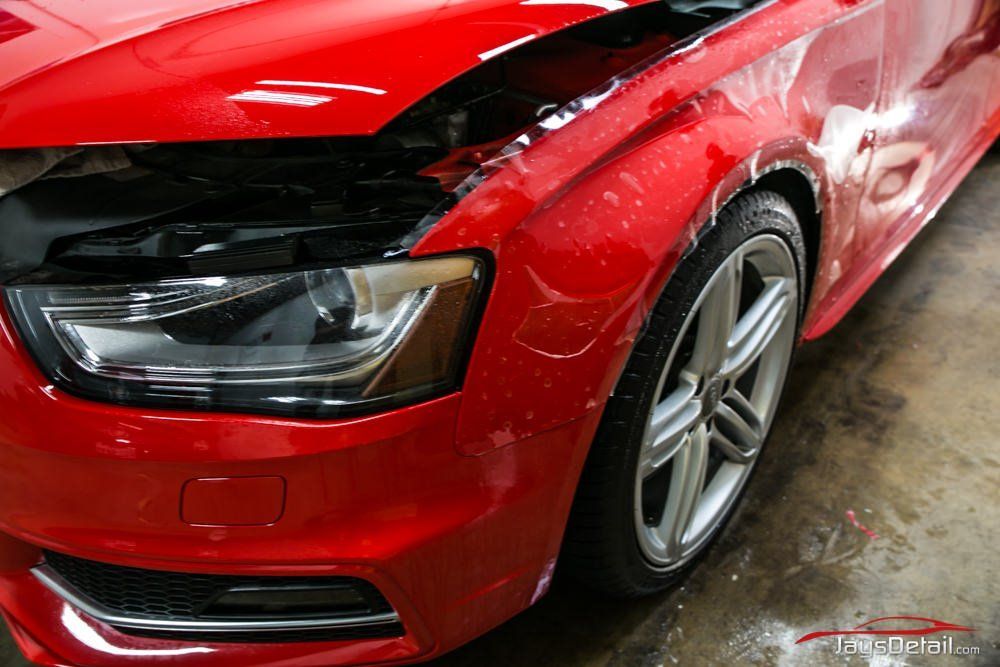 Red car with hood open, partially covered in clear film, showing a headlight and wheel.