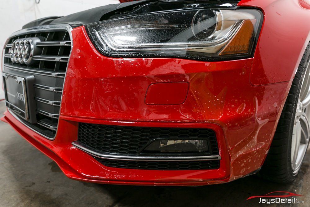 Red Audi car bumper with clear protective film, showing grill, headlight, and chrome trim.