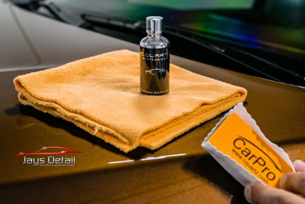 A car detailing setup: bottle of product, orange microfiber cloth, applicator on a car's hood.
