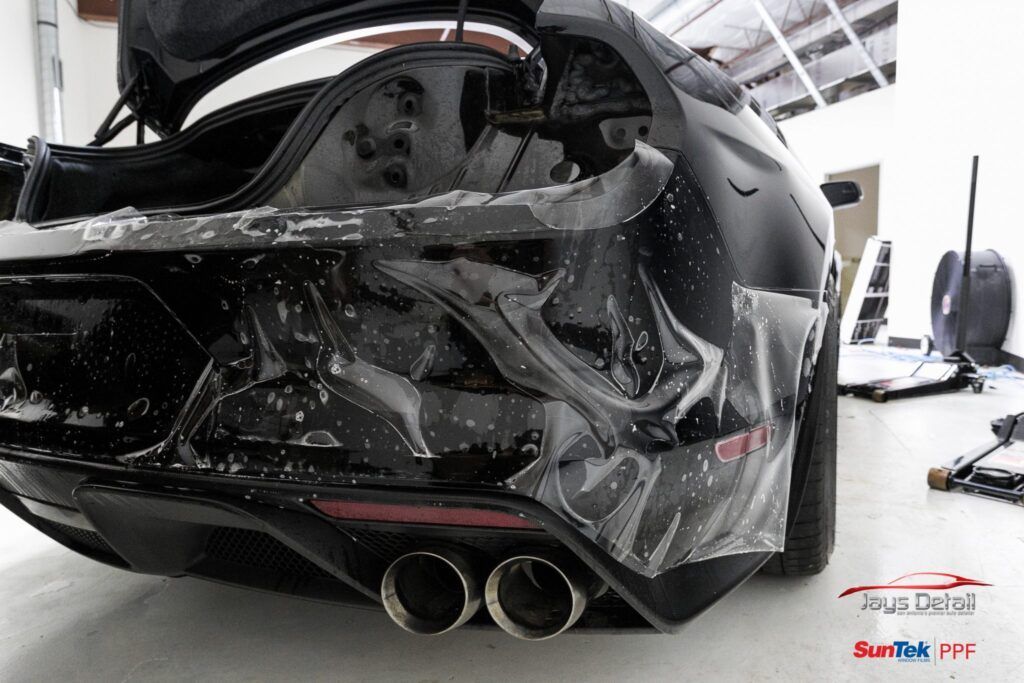 Black car bumper with protective film being applied; silver exhaust tips.