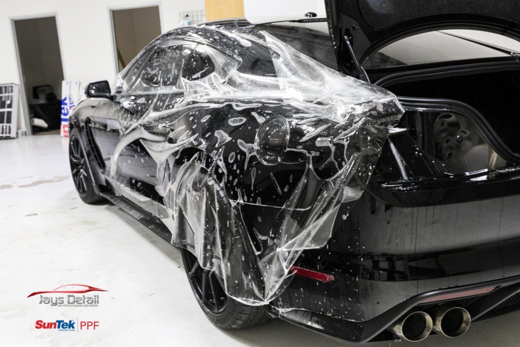 Black car with protective film being applied in a workshop, trunk open.