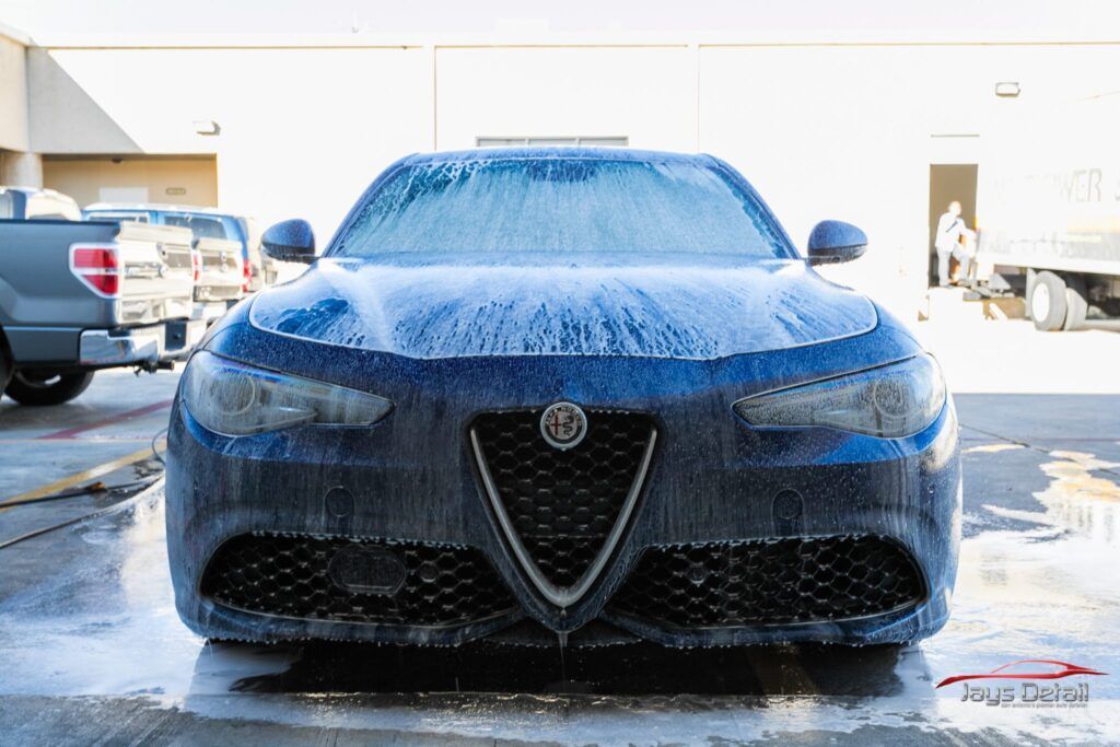 Blue Alfa Romeo being washed, covered in soapy foam. Front view, outdoor setting.