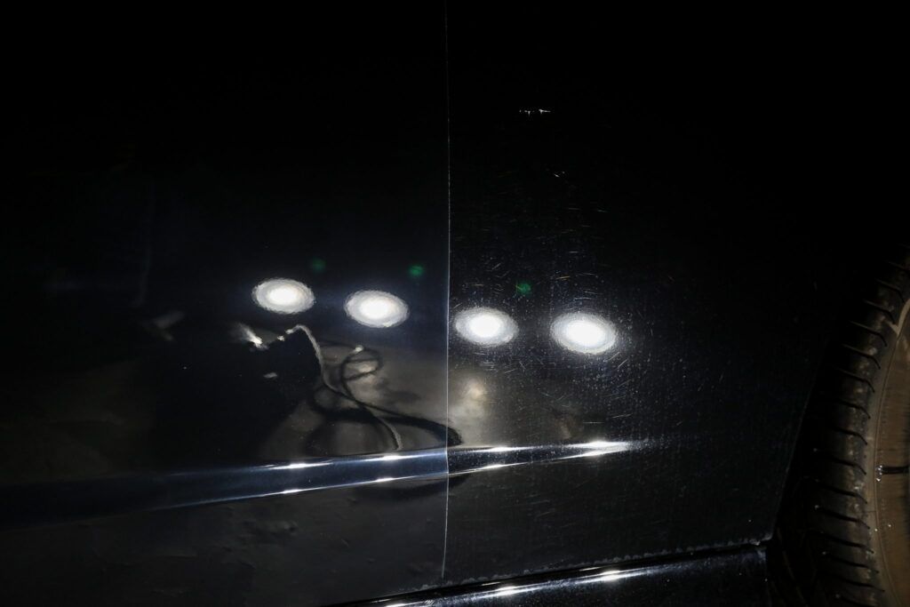 Black car door reflecting bright lights, with a visible scratch.