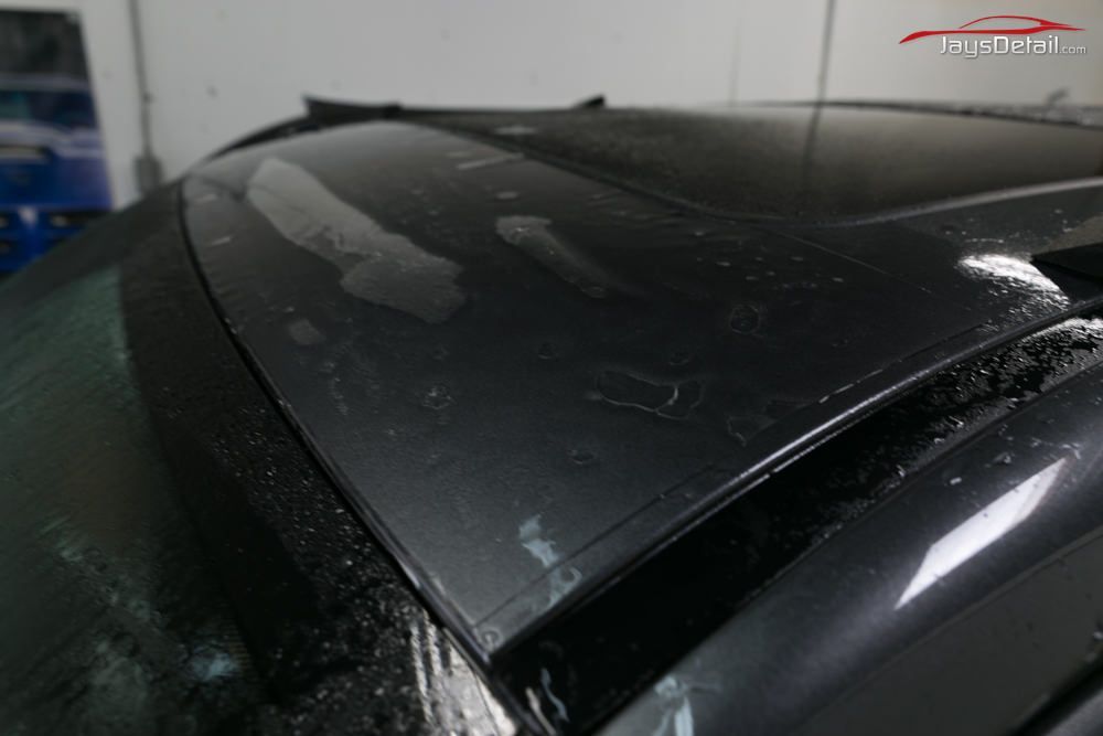 Close-up of a dark car roof with damaged protective film peeling off, likely in a garage setting.