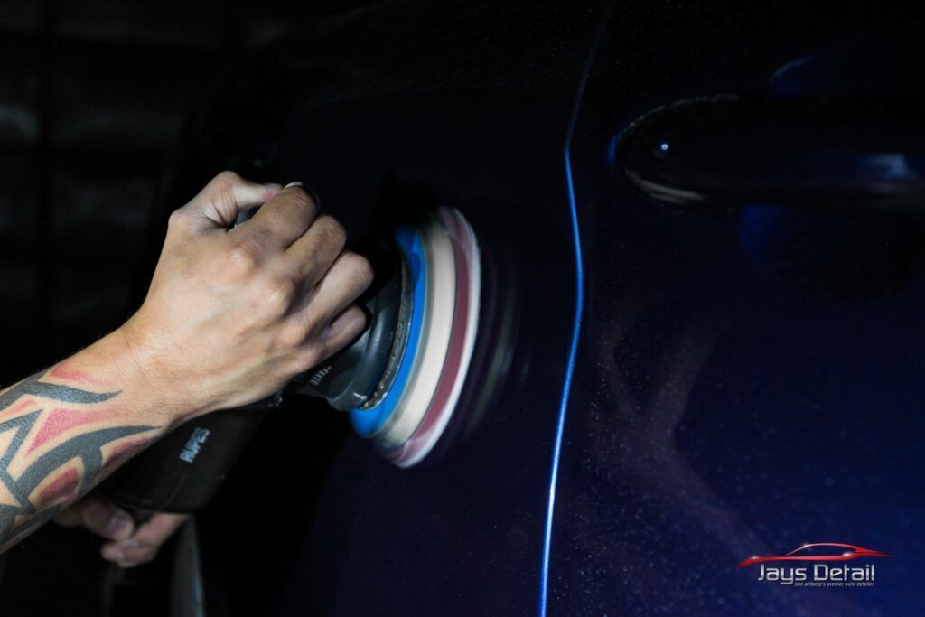 Person polishing a dark blue car door with a rotating buffer, indoors.