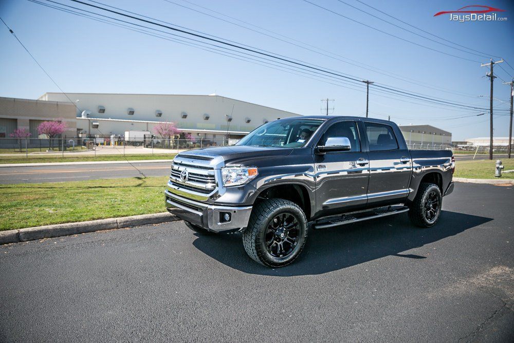 Dark gray Toyota Tundra truck parked on pavement beside green grass and a building on a sunny day.