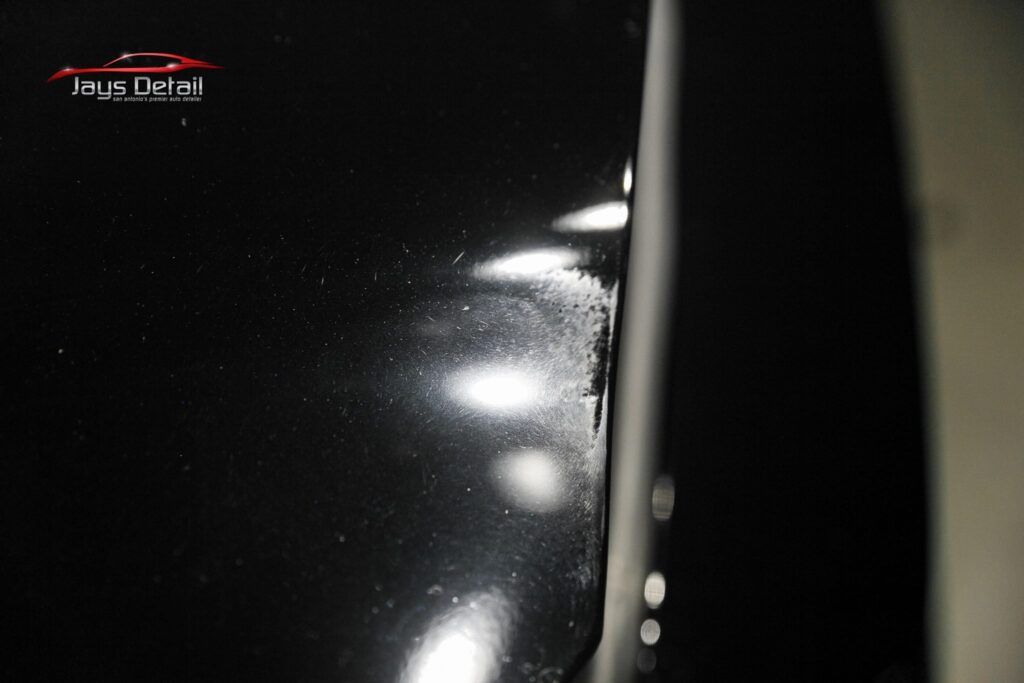 Close-up of a black car panel with scratches and visible damage near an edge, light reflecting off the surface.