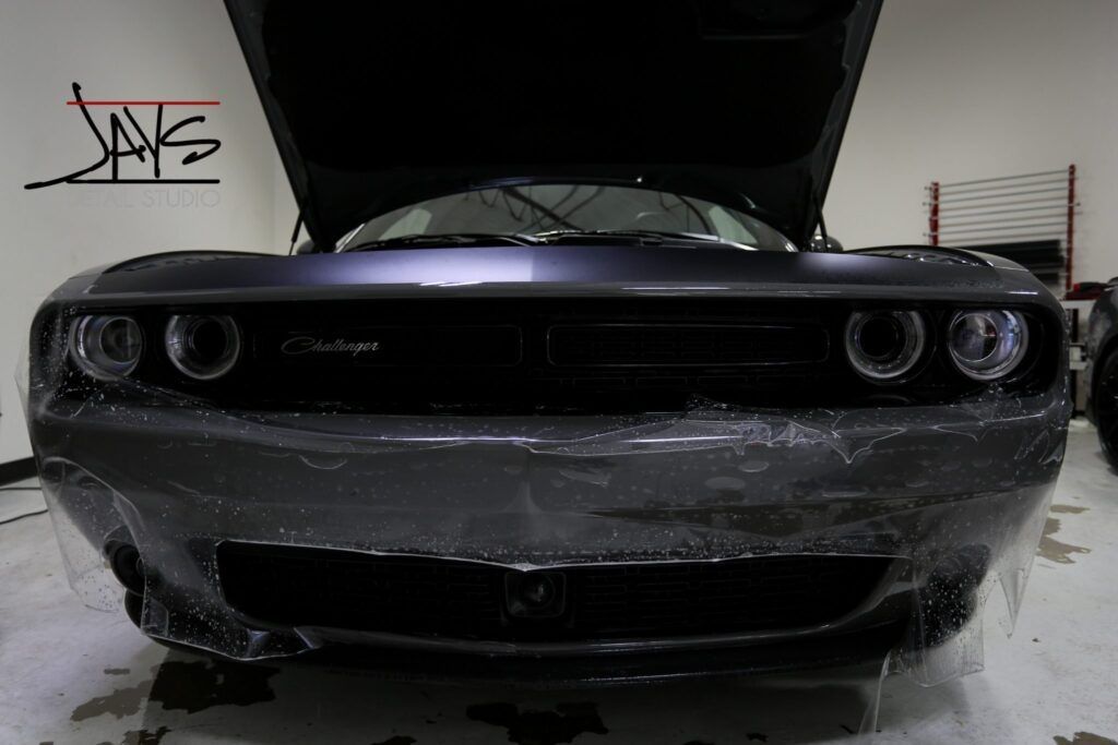A dark gray Dodge Challenger with hood open, covered in clear protective film, in a shop.